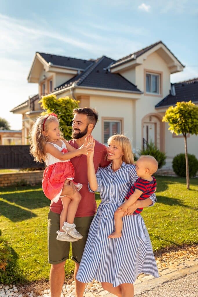 Happy family of four standing in front of a suburban house on a sunny day, with parents holding their young children.