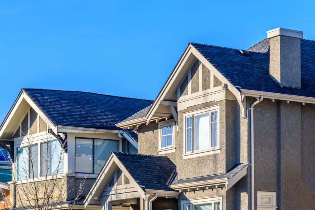 Upper exterior view of modern stucco homes featuring dark shingled roofs, gable rooflines, and white-framed windows under a clear blue sky.